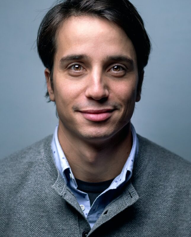 A man in a gray sweater and collard shirt faces the camera for a portrait.