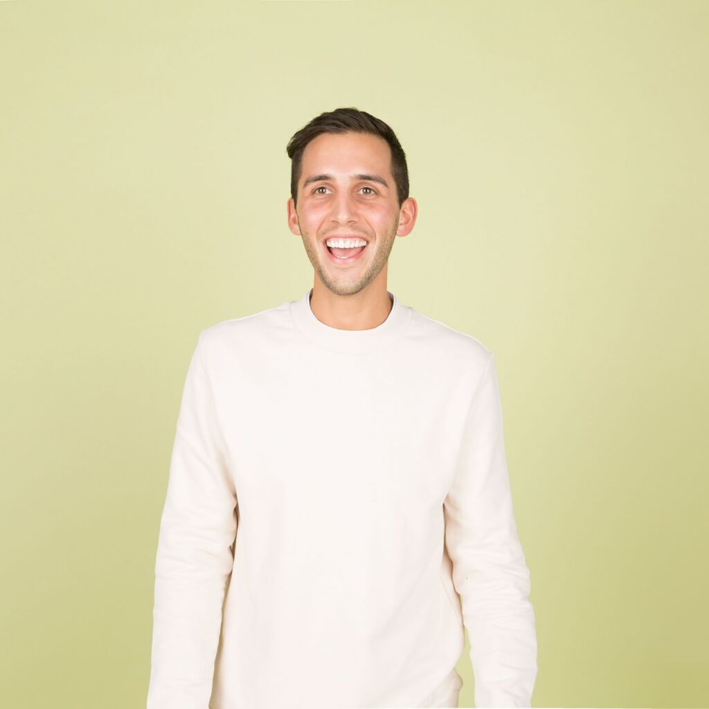 A man in a white sweater stands against a yellow backdrop.