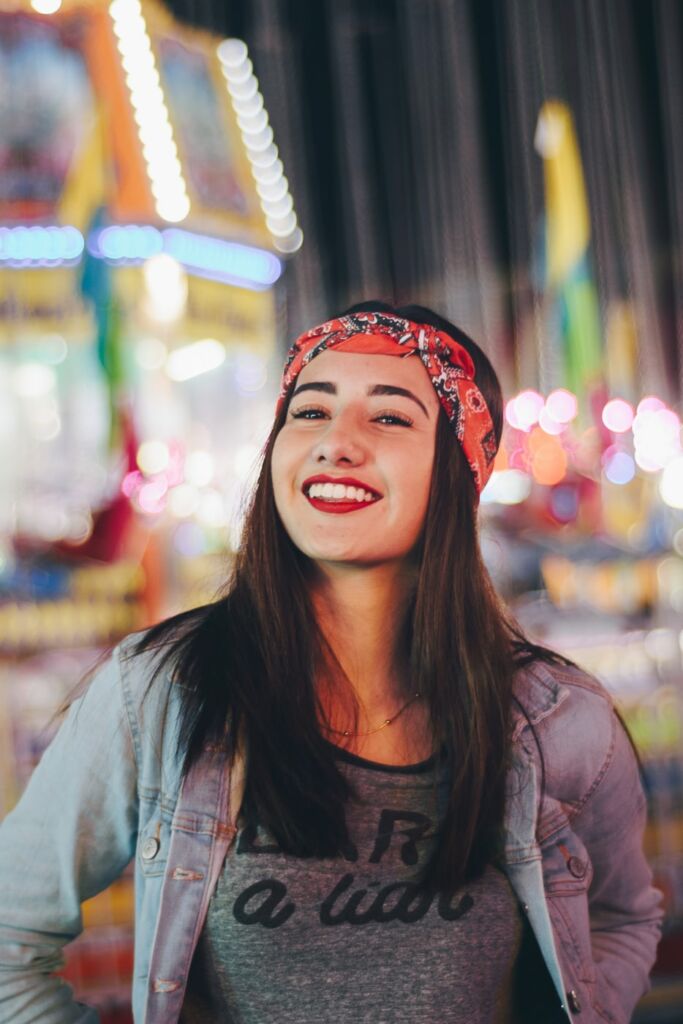 A woman in a jeans jacket, headband, and red lipstick smiles in front of lights.